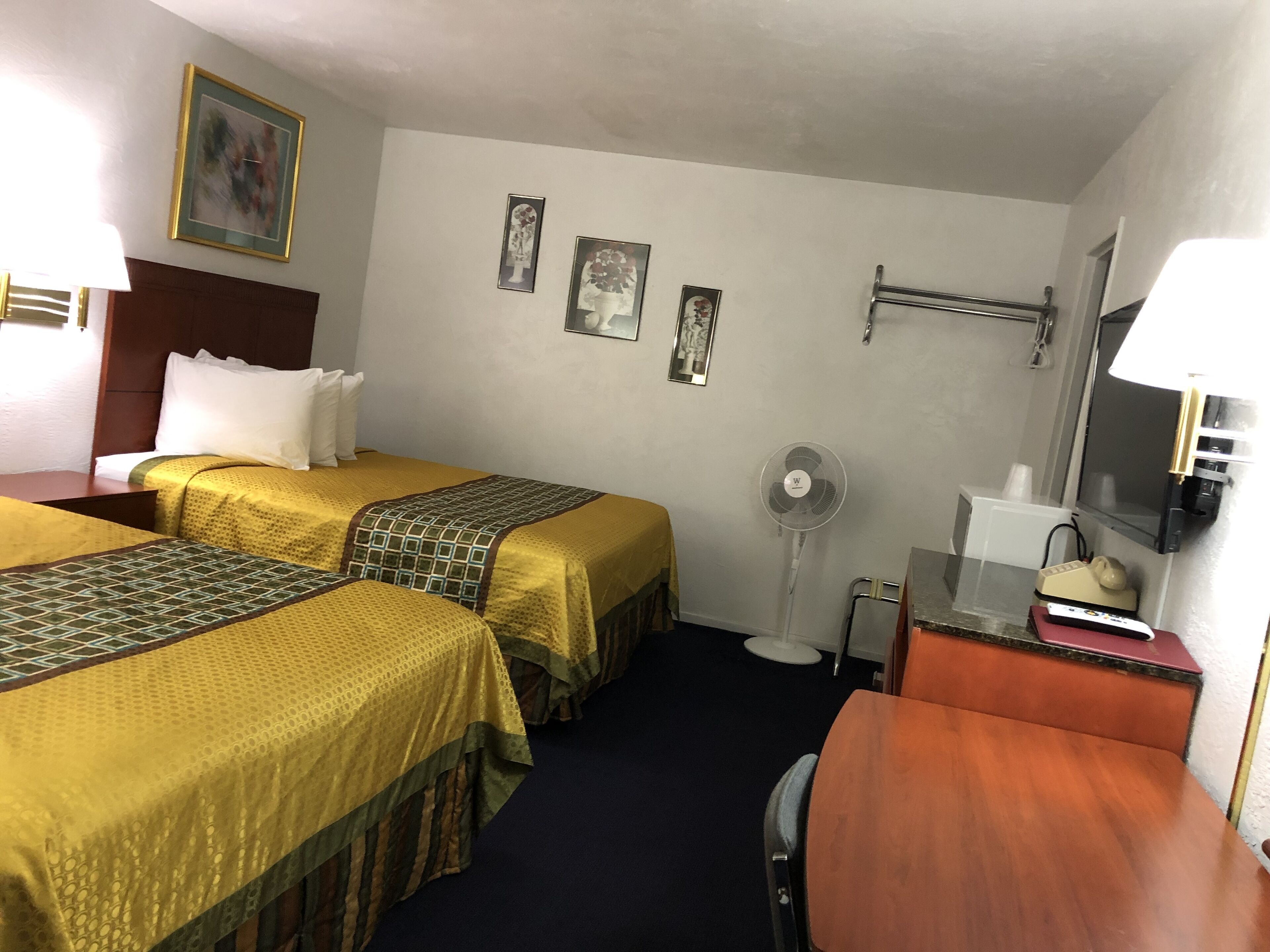 A hotel room with two beds, a desk, a chair, a fan, a TV, and a small table with a lamp.