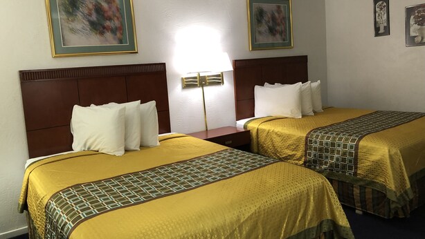 A hotel room with two beds, each with a headboard and a patterned bedspread. There are framed artworks on the walls and a bedside lamp between the beds.