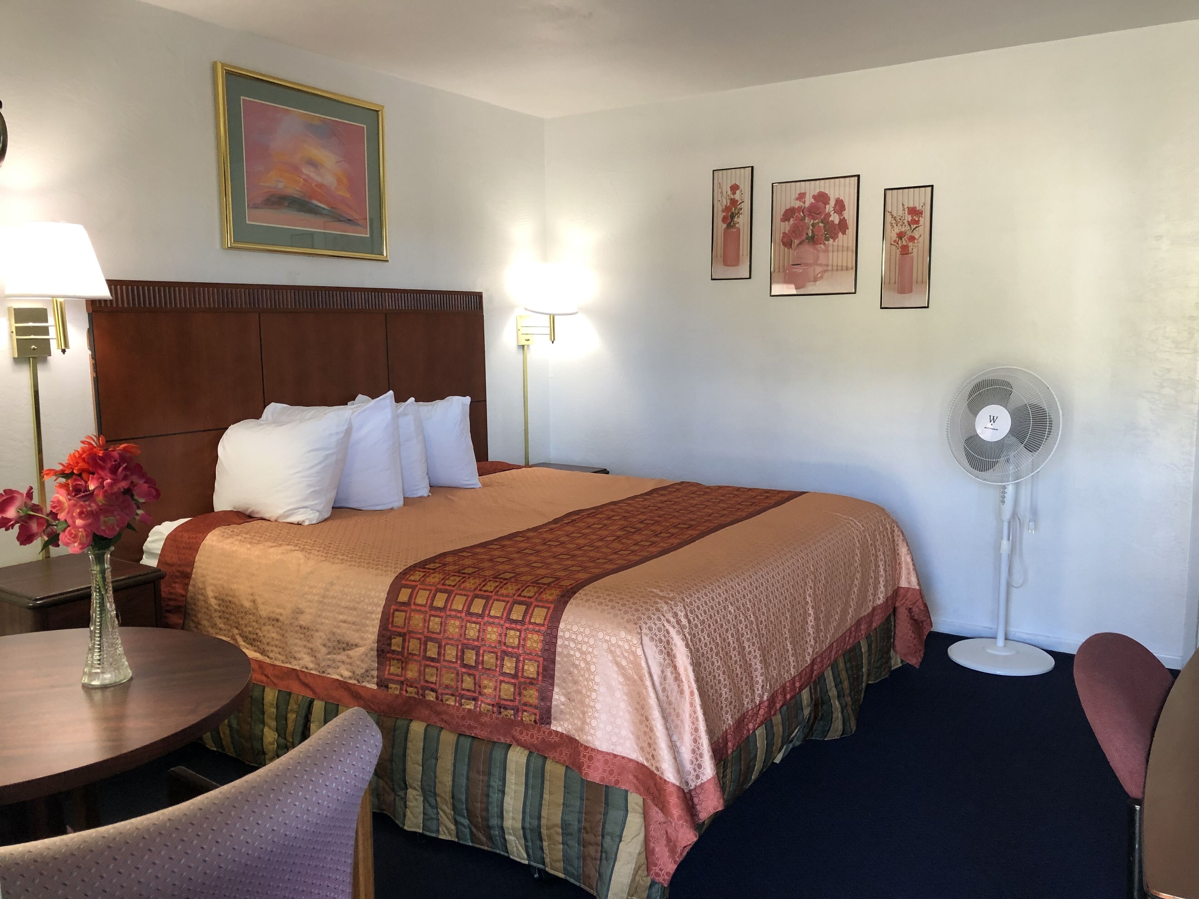 A hotel room with a bed, bedside table, lamp, chair, and wall-mounted fan.