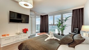 A modern hotel room with a large bed, a flat-screen TV, a potted plant, and a view of the city through large windows. - Apartinfo Apartments - Sadowa (Gdansk)