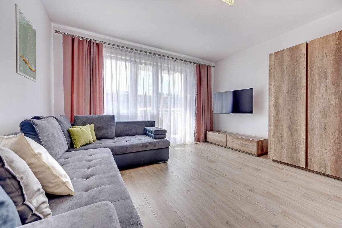 Uma sala de estar moderna com um sofá cinzento, piso de madeira, uma TV fixada na parede e um guarda-roupa de madeira grande.