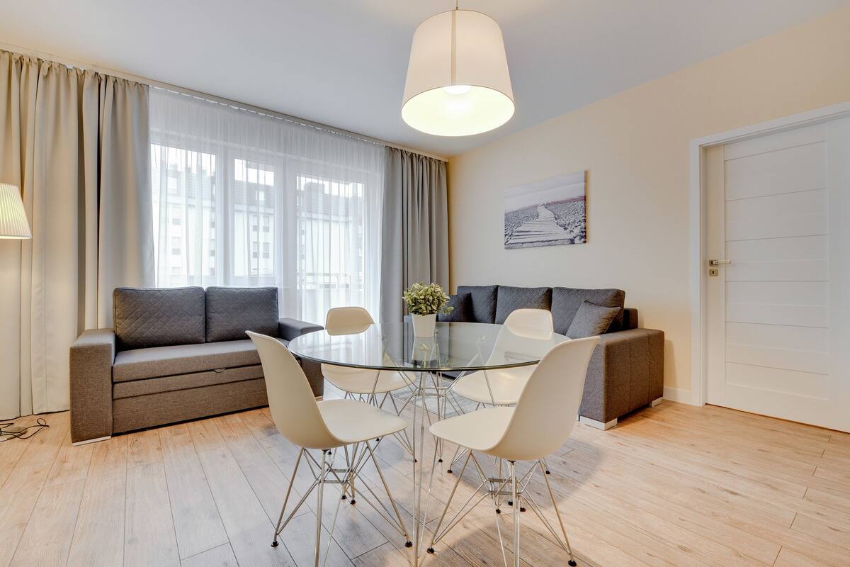 Uma sala de estar moderna com uma mesa de jantar de vidro cercada por cadeiras, um sofá e uma janela grande com cortinas.