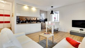 A modern living room with a white sofa, a coffee table, and a red pillow. There is a dining area with a white table and chairs, and a kitchen with white cabinets and a black countertop. A TV is mounted on the wall, and a window provides natural light. - Apartinfo Apartments - Sadowa (Gdansk)