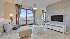 A modern living room with a sofa, a round coffee table, a TV on a brick wall, and large windows with curtains. - Apartinfo Island Apartments (Gdansk)