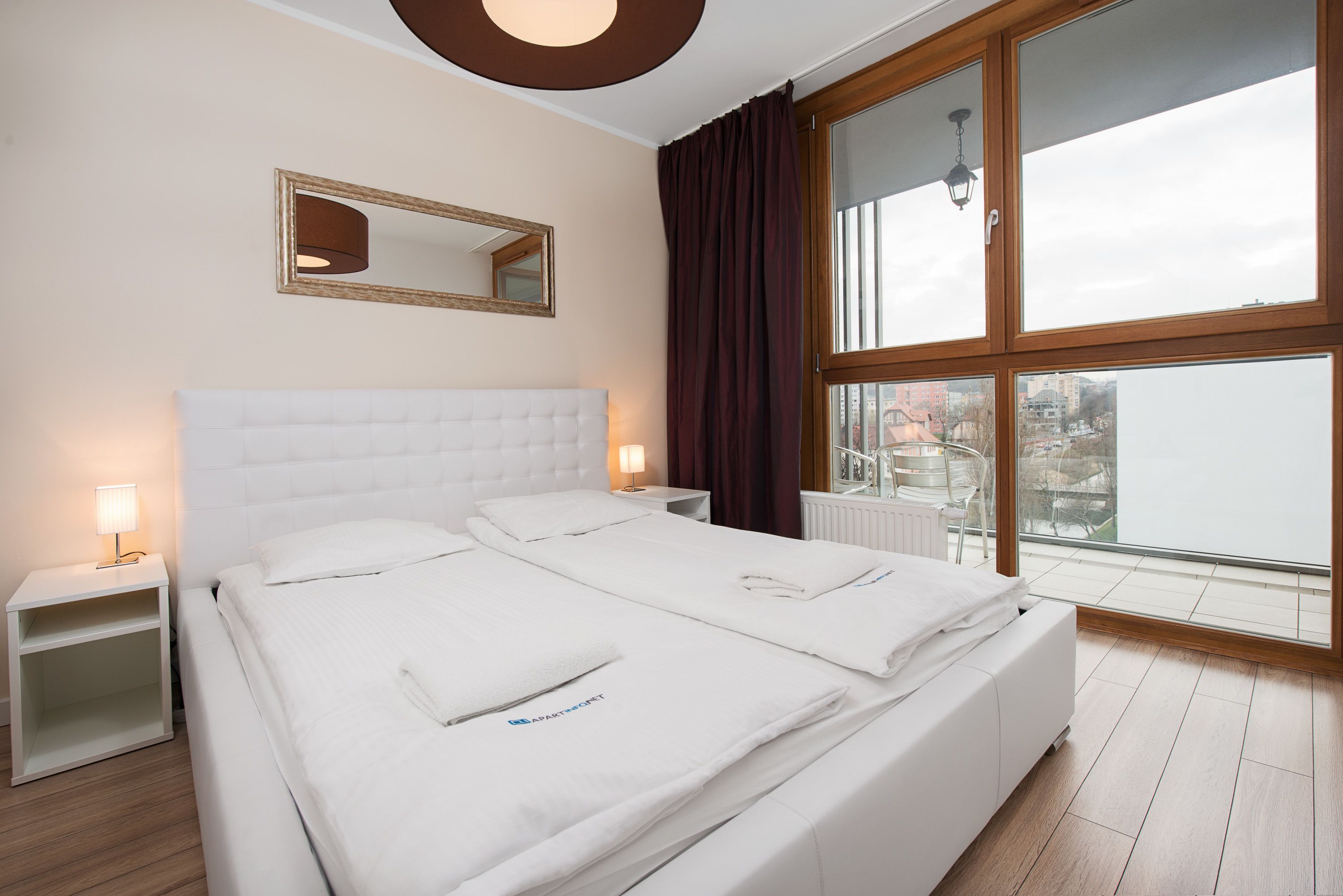 Ein Hotelzimmer mit einem großen Bett, einem Nachttisch, einem Spiegel und einem Fenster mit Blick auf Gebäude.