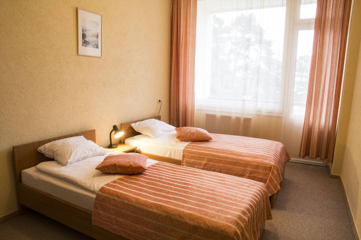 Quarto com duas camas, mesa de cabeceira, abajur e janela com cortinas.