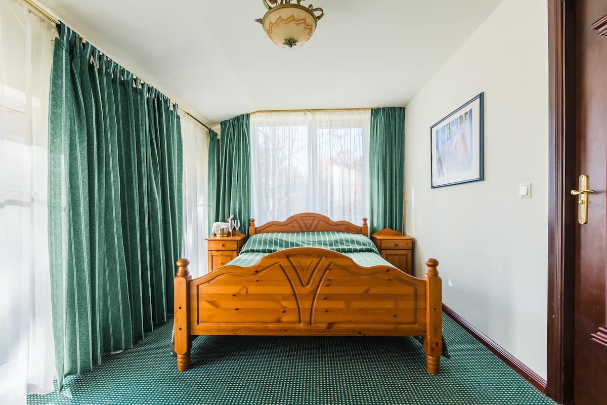 Uma cama de madeira com colcha verde e branca listrada, duas mesas de cabeceira, uma mesinha de apoio, uma janela com cortinas translúcidas e um quadro na parede.