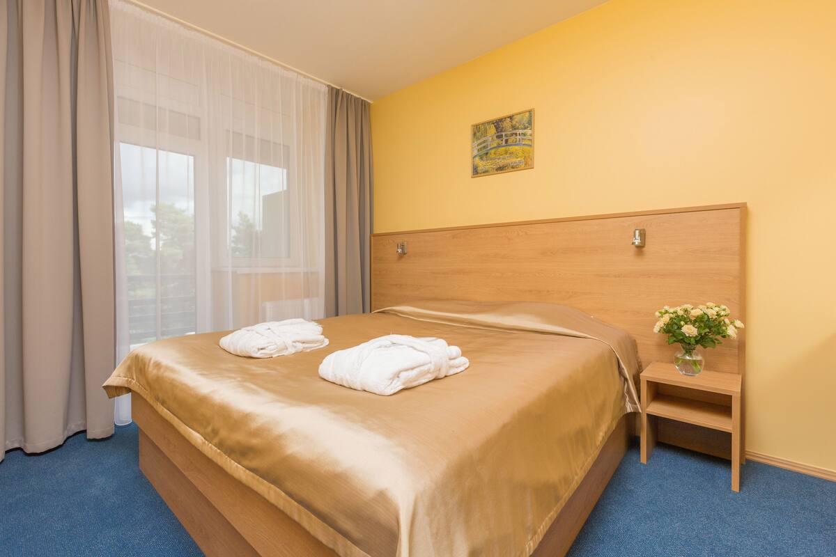 Quarto de hotel com uma cama grande, mesa de cabeceira com um vaso de flores e um quadro na parede.