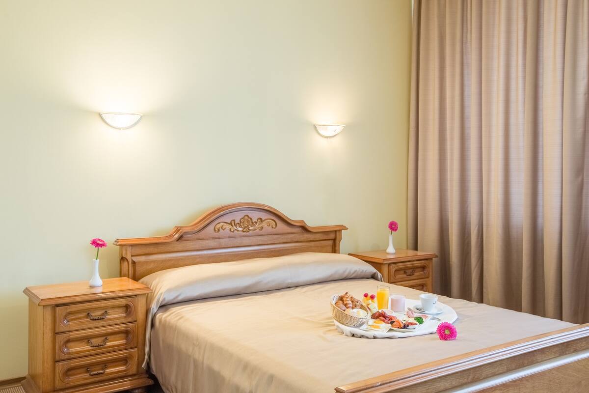 Cama arrumada com uma bandeja de itens de café da manhã, duas luminárias de cabeceira e uma mesinha de cabeceira de madeira com gavetas.