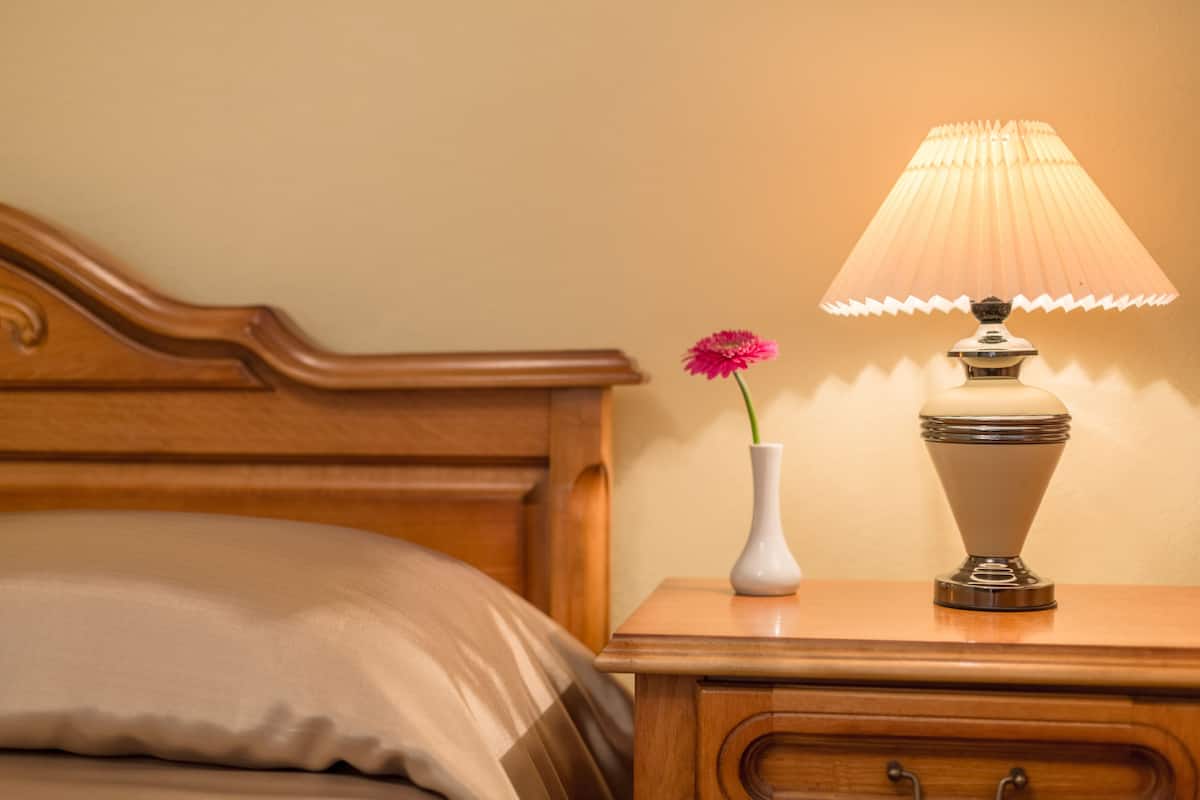 Uma mesa de cabeceira de madeira com uma luminária e um vaso com uma flor rosa.