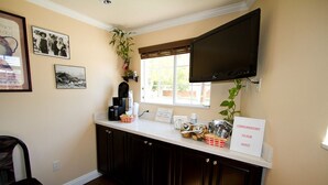 A kitchen with dark cabinets, a white countertop, a television mounted above it, and a window with blinds. - Trails Motel (Lone Pine)
