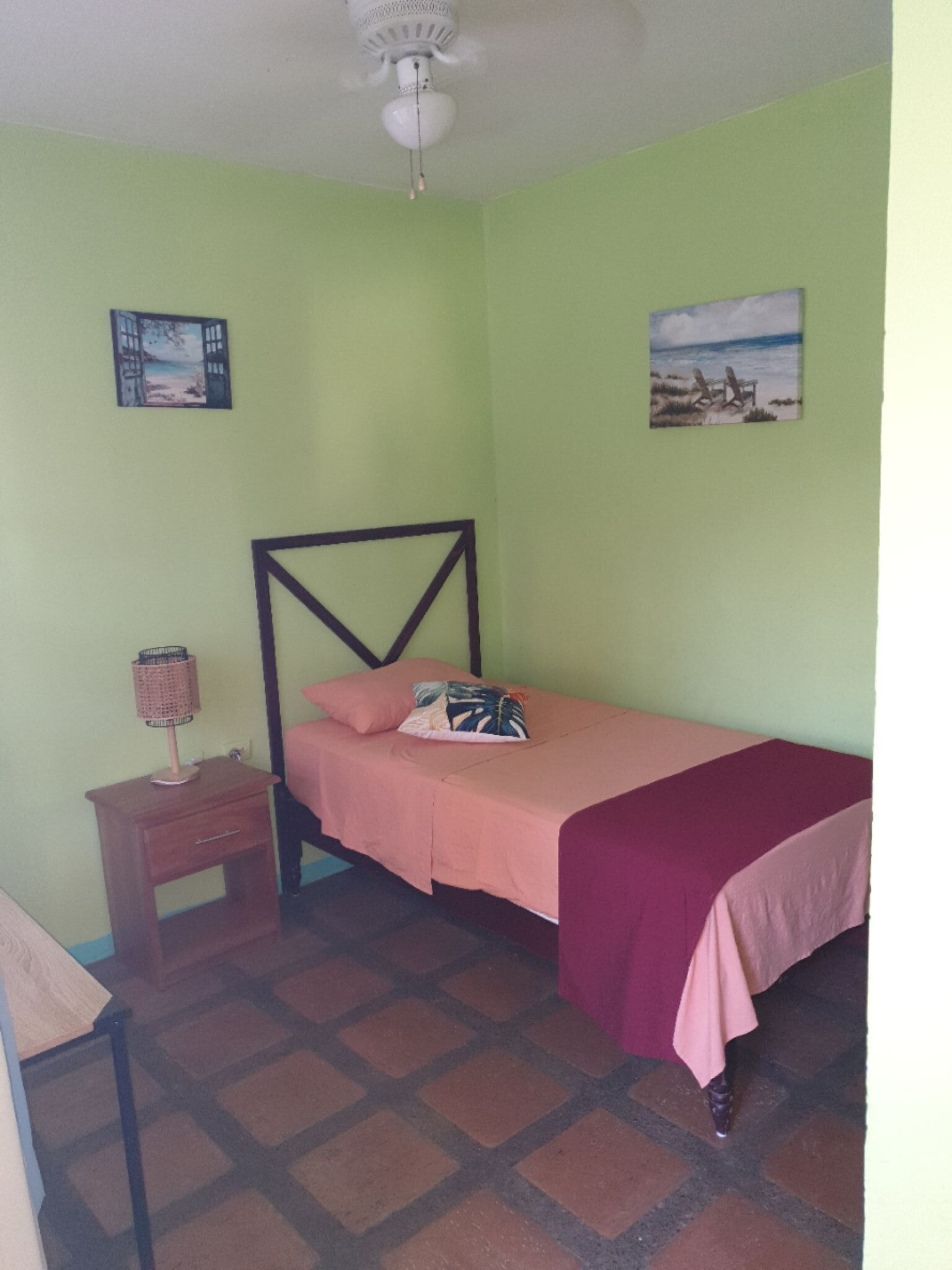 Un dormitorio con una cama individual, una mesita de noche, un ventilador de techo y dos obras de arte en la pared.