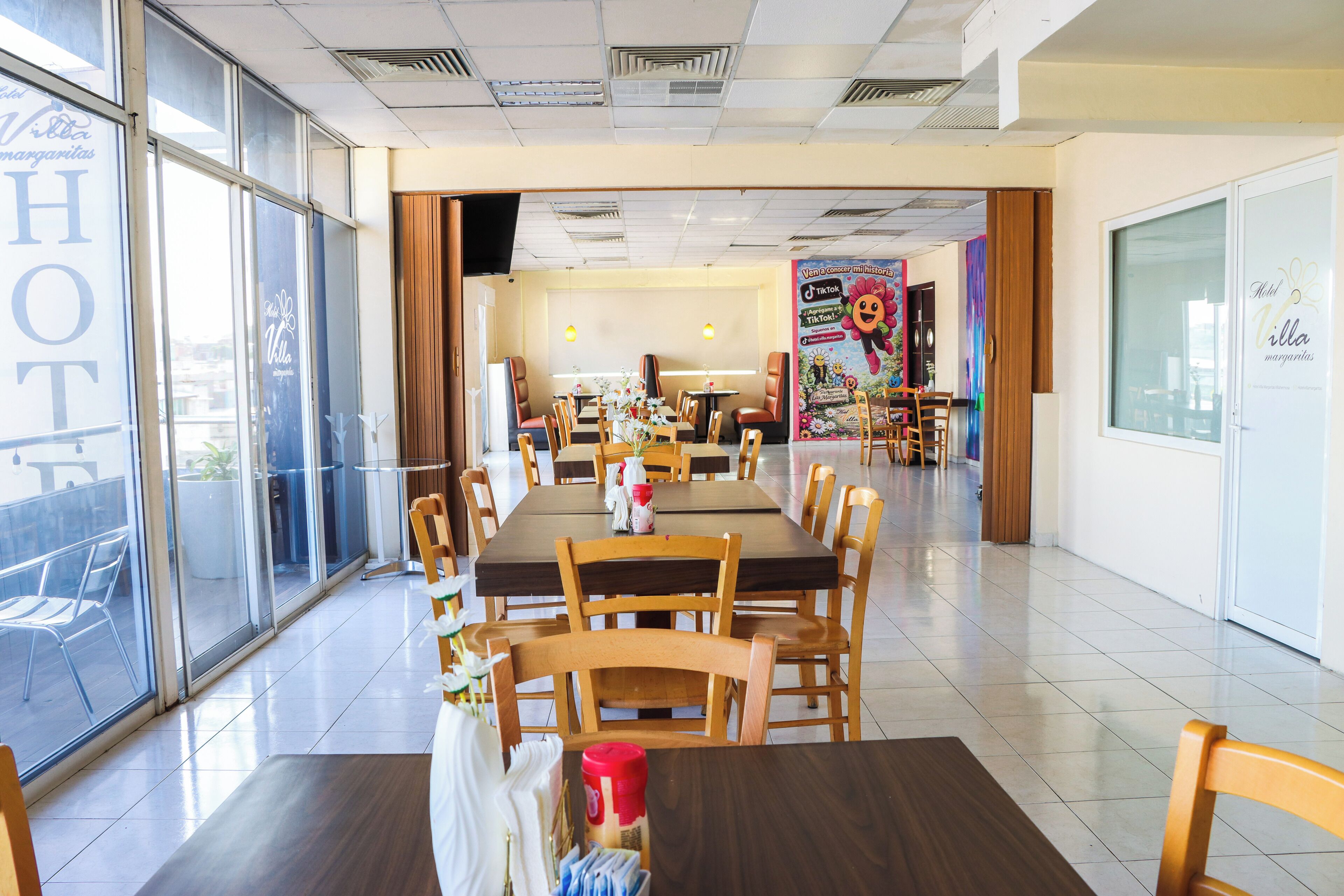 A hotel dining area with multiple tables and chairs, large windows, and a view of the outside.