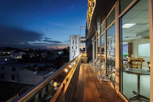 Un balcón de hotel con vistas a la ciudad al atardecer, con barandilla, sillas y un cartel con el nombre del hotel.