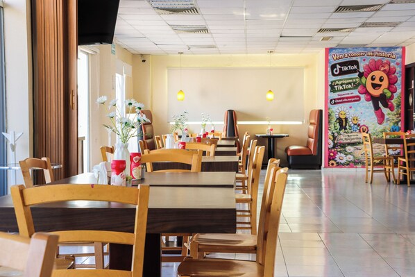 A restaurant with wooden tables and chairs, a flower vase on each table, and a wall with a colorful poster.