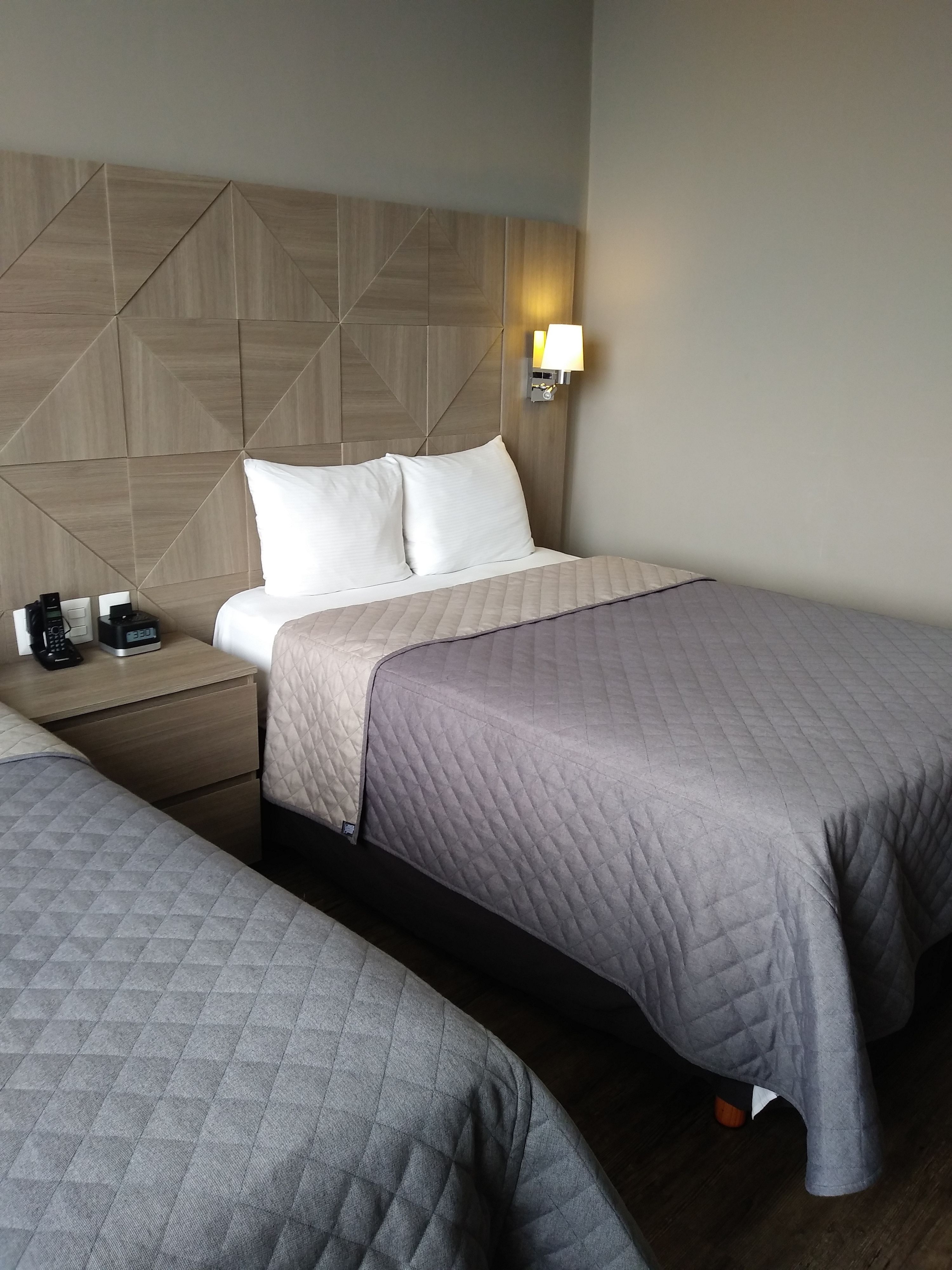 Una habitación de hotel con dos camas, un cabecero de madera, una mesita de noche con una lámpara y un mueble auxiliar con un teléfono y una cafetera.