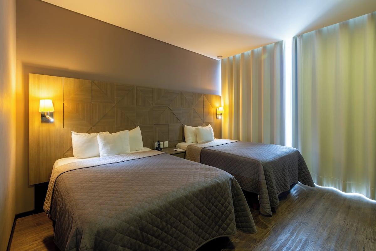 Habitación de hotel con dos camas, cabecero de madera, lámparas de noche y un ventanal con cortinas.