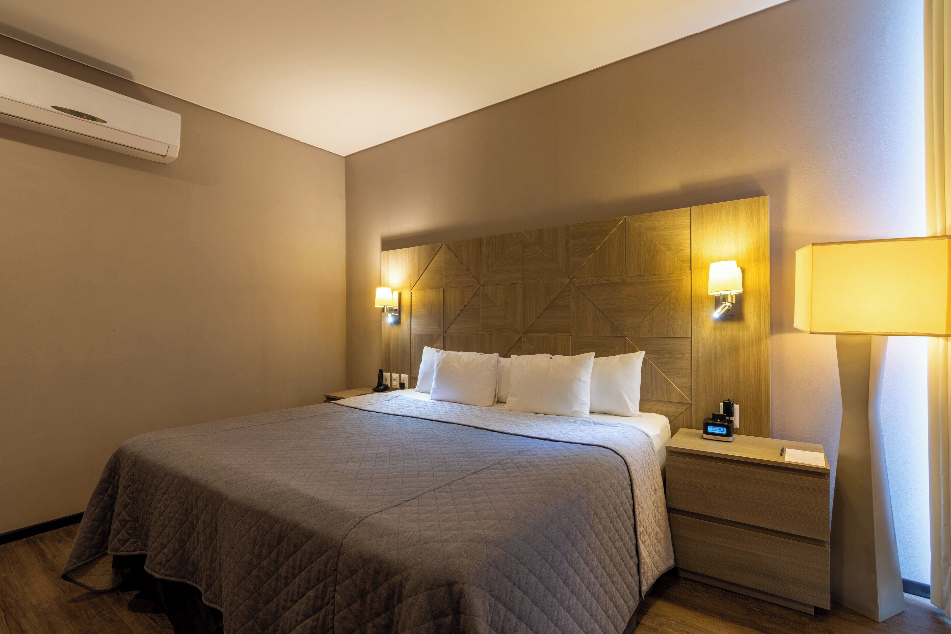 Een hotelkamer met een groot bed, twee nachtkastjes met lampen, een houten hoofdbord en een airconditioning aan de muur.