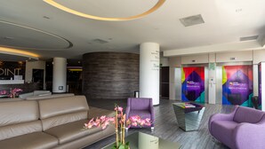 A modern hotel lobby with a reception desk, a seating area with a sofa and armchairs, a coffee table with a vase of flowers, and a colorful wall with the hotel's logo. - Hotel Glow Point - Mulza (León)