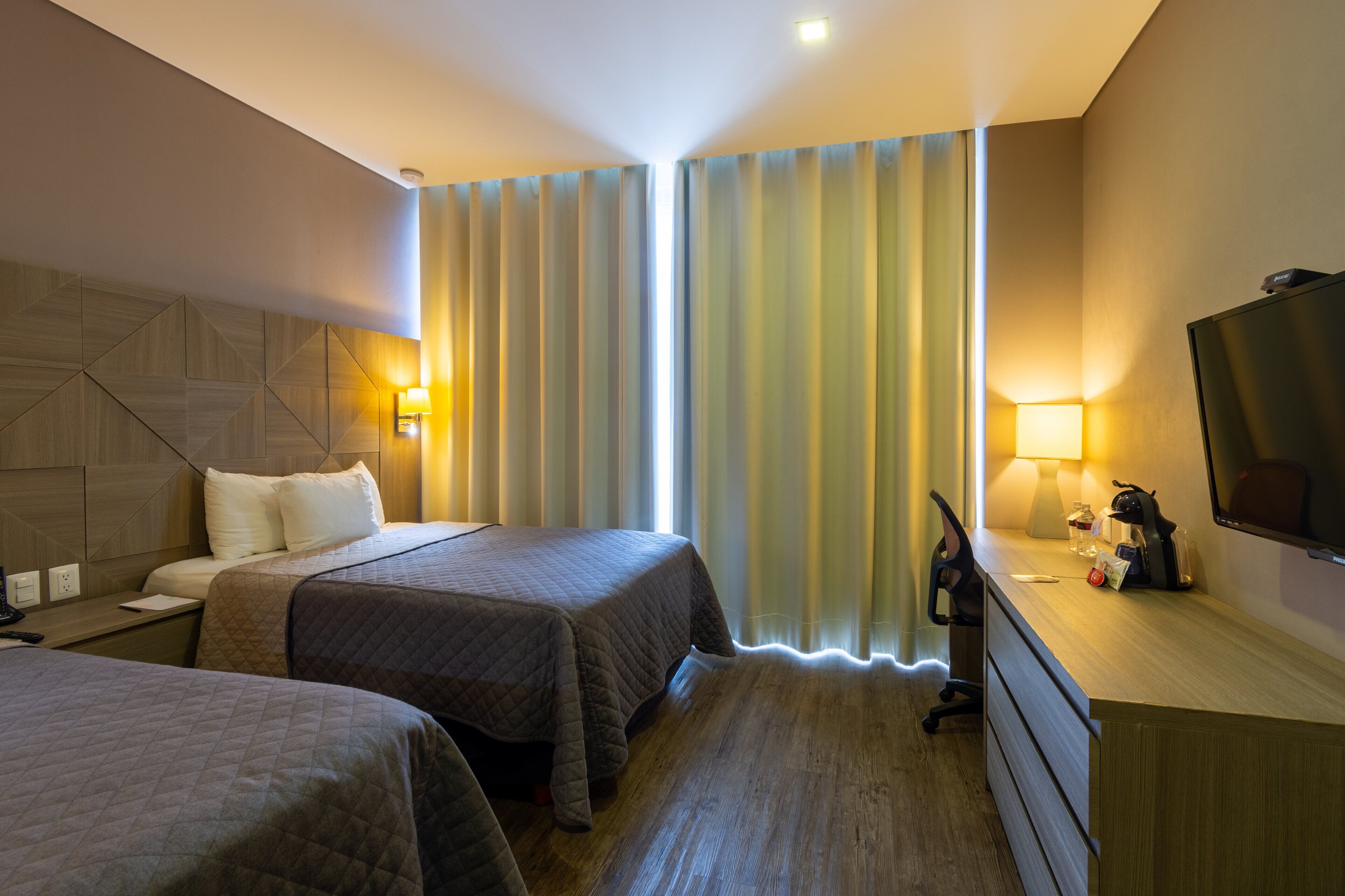 Habitación de hotel con dos camas, un escritorio con lámpara, televisión y ventana con cortinas.
