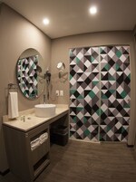 A modern bathroom with a round mirror, a sink, and a shower with a geometric-patterned door.