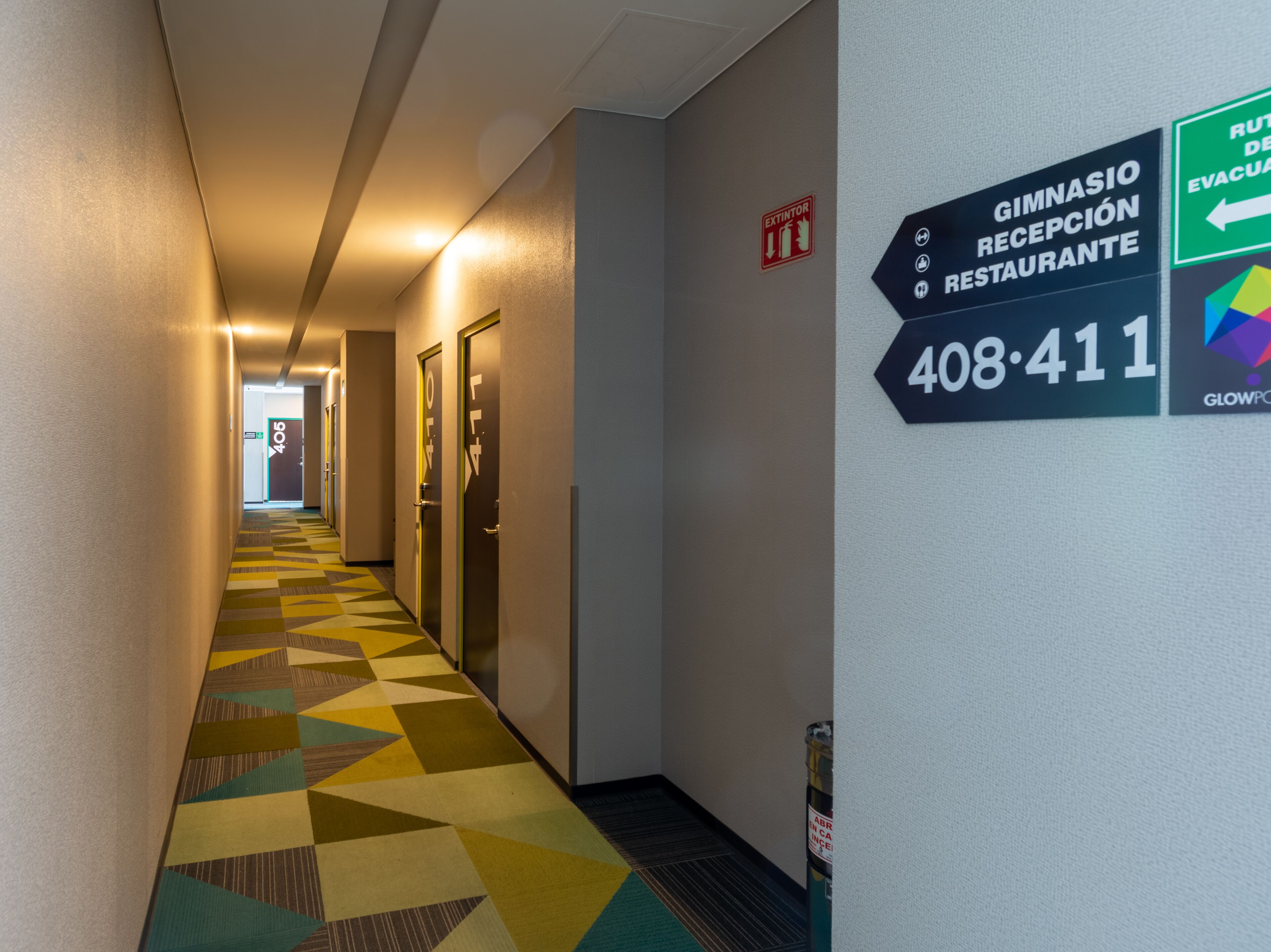 Un pasillo de hotel con alfombra de diseño geométrico, puertas numeradas 406 y 411, e indicaciones para un gimnasio, recepción y restaurante.