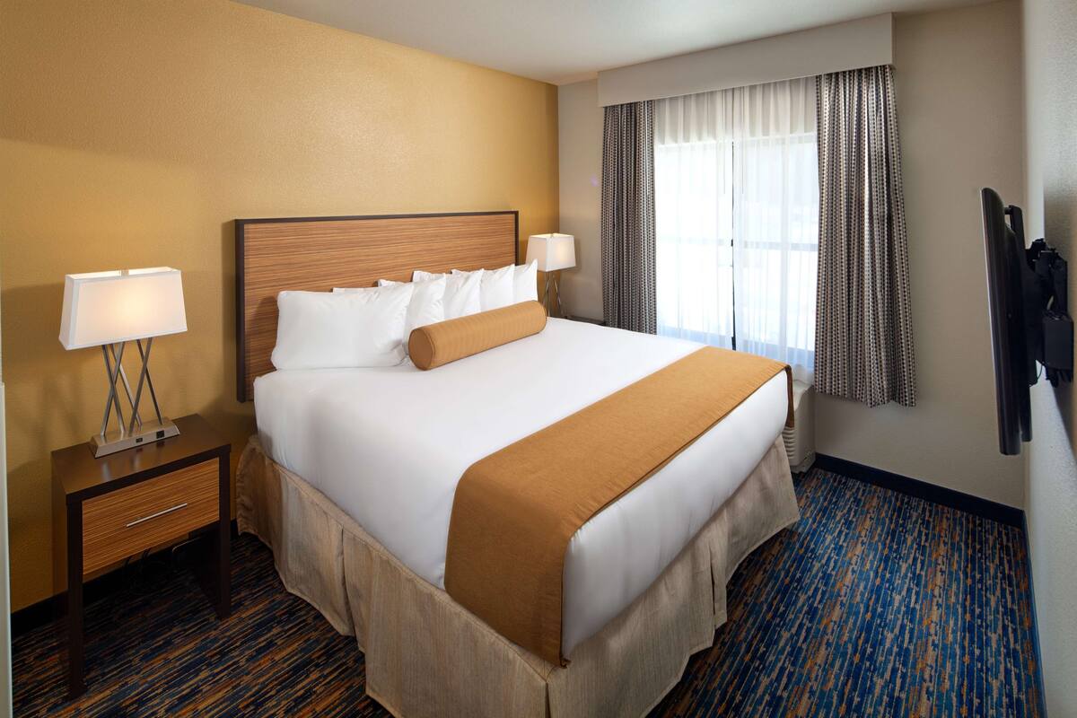 A hotel room with a large bed, two bedside tables with lamps, a window with curtains, and a coat rack.