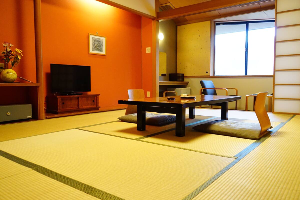 Ein Raum mit Tatami-Matten, einem niedrigen Tisch, Stühlen, einem Fernseher und einer Vase mit Blumen.