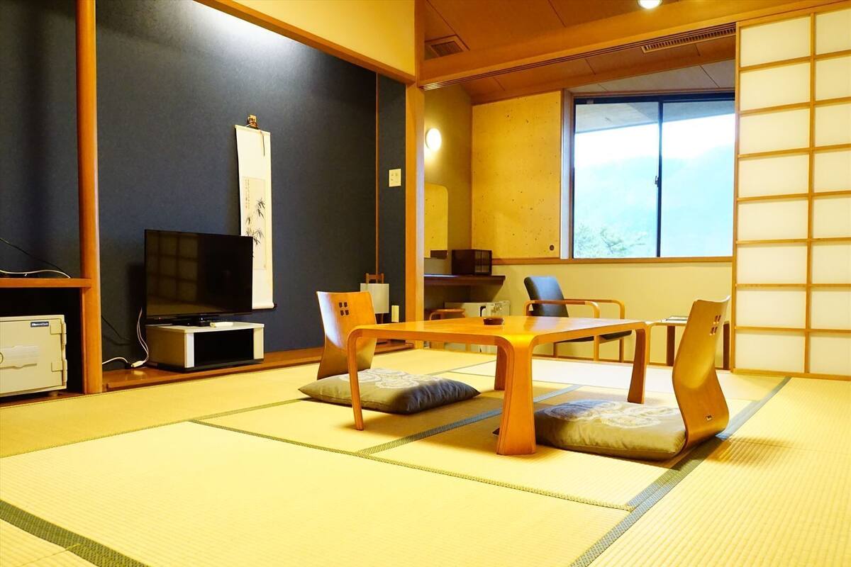 Ein Raum mit Tatami-Matten, einem niedrigen Tisch, Stühlen, einem Fernseher und einer Schiebetür.