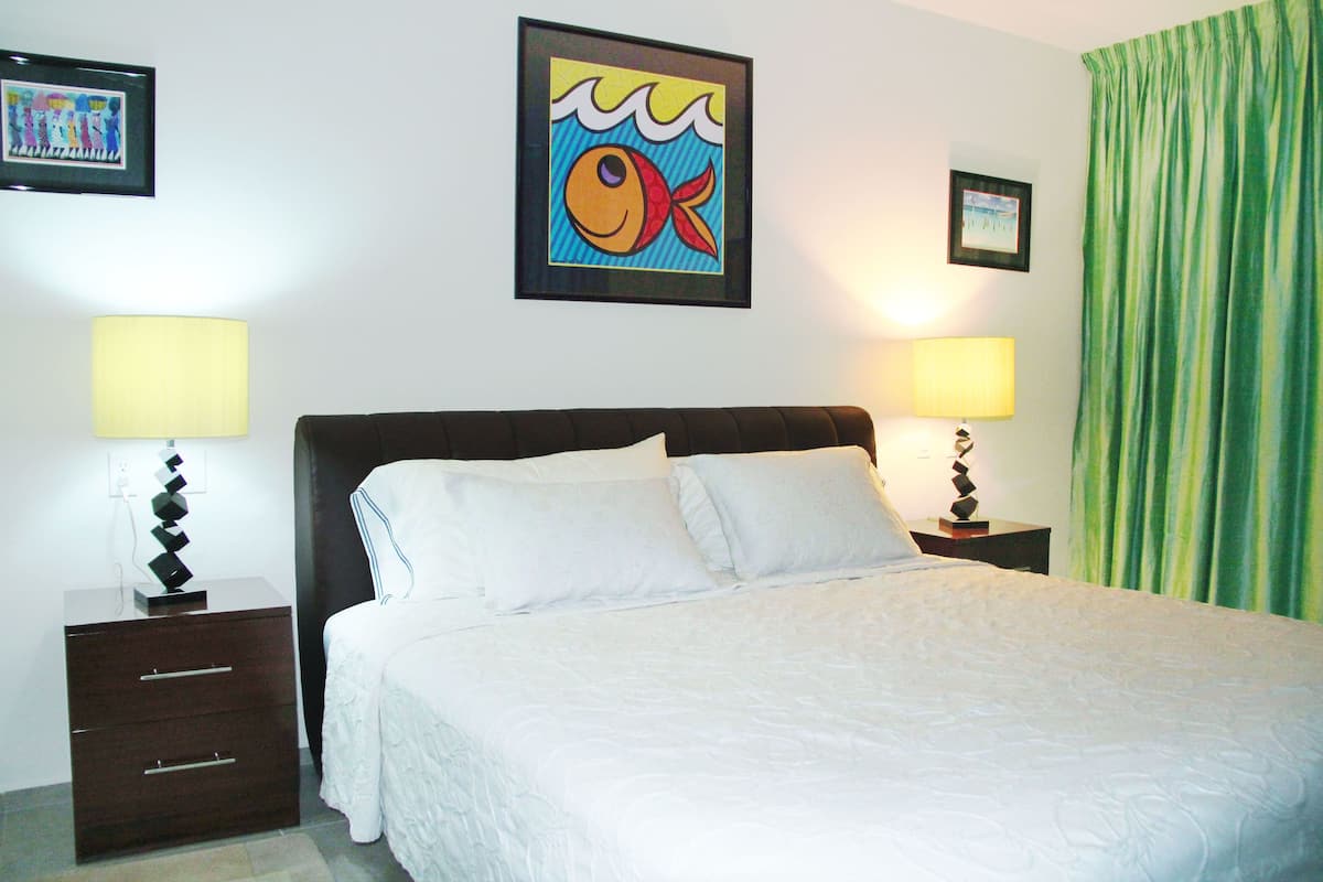 A neatly made bed with white linens, two bedside tables with lamps, a framed artwork above the bed, and a green curtain to the right.