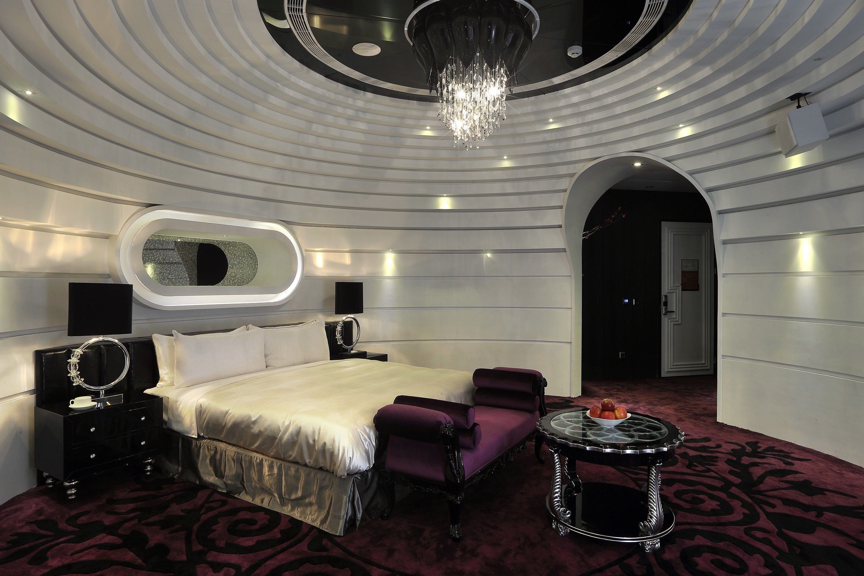 A modern hotel room with a large bed, a vanity with a mirror, a round table with a fruit bowl, and a chandelier.