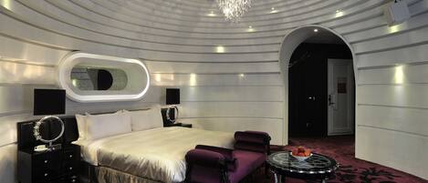 Une chambre d’hôtel moderne avec un grand lit, une coiffeuse avec un miroir, une table ronde sur laquelle se trouve un panier de fruits, et un lustre.