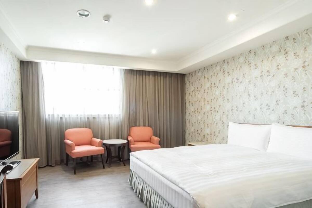 A hotel room with a bed, two chairs, a small table, a TV, and a window with curtains.