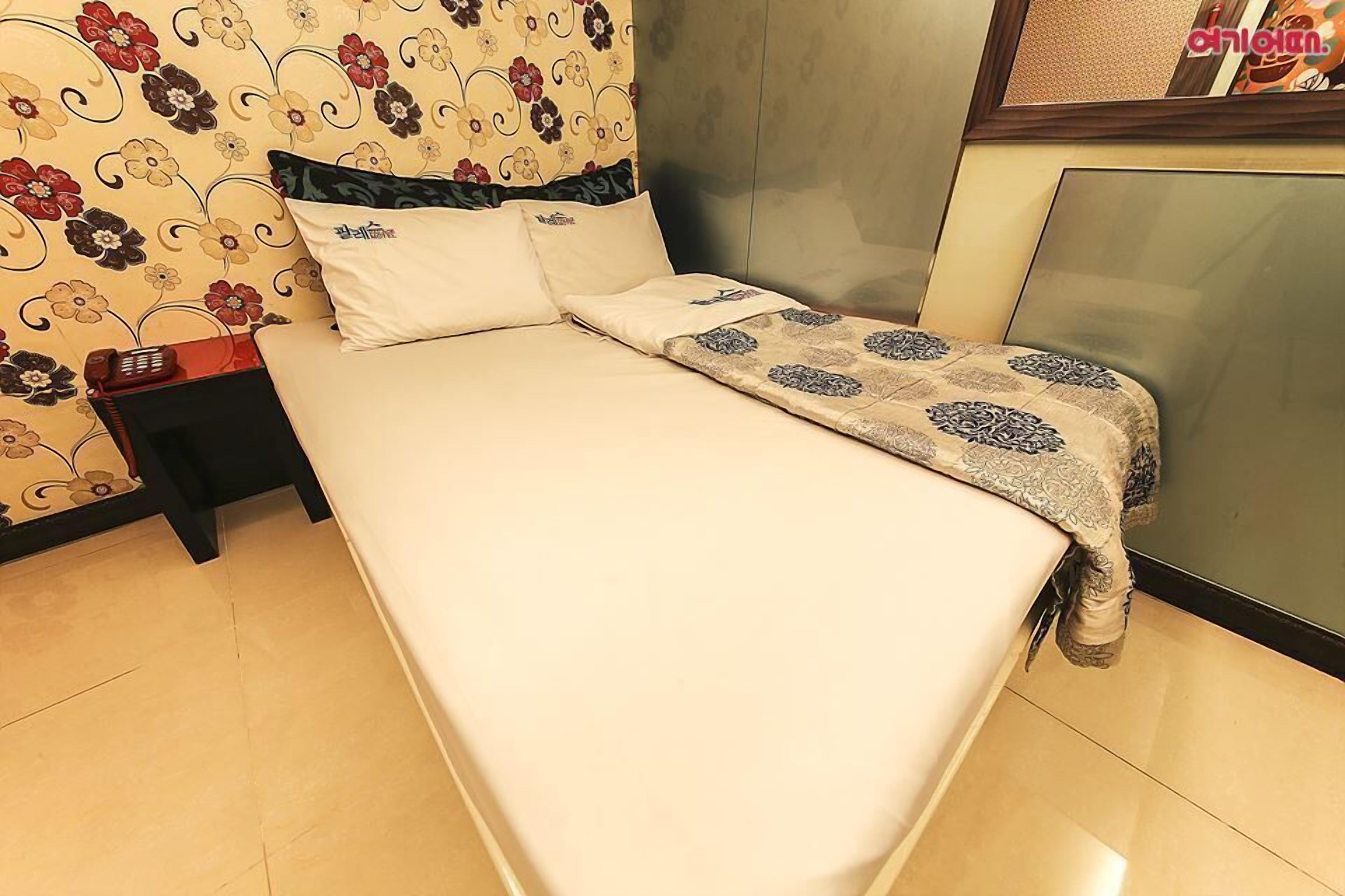A neatly made bed with a patterned bedspread, a bedside table with a phone, and a floral wallpaper.