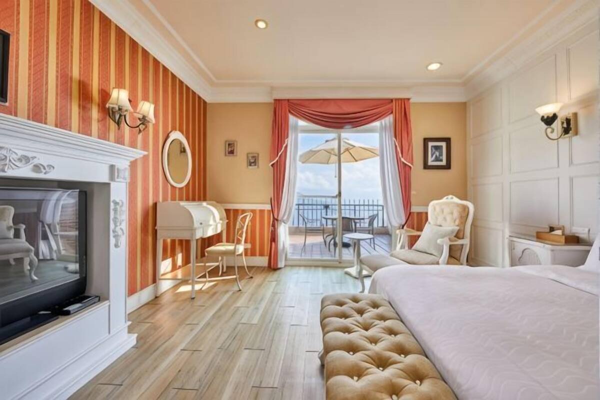 Une chambre d’hôtel équipée d’une télévision, d’une cheminée, d’un balcon avec une table et des chaises, et d’un grand lit.