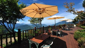Un patio al aire libre con mesas y sillas de madera, sombrillas color beige y una vista panorámica de la montaña.
