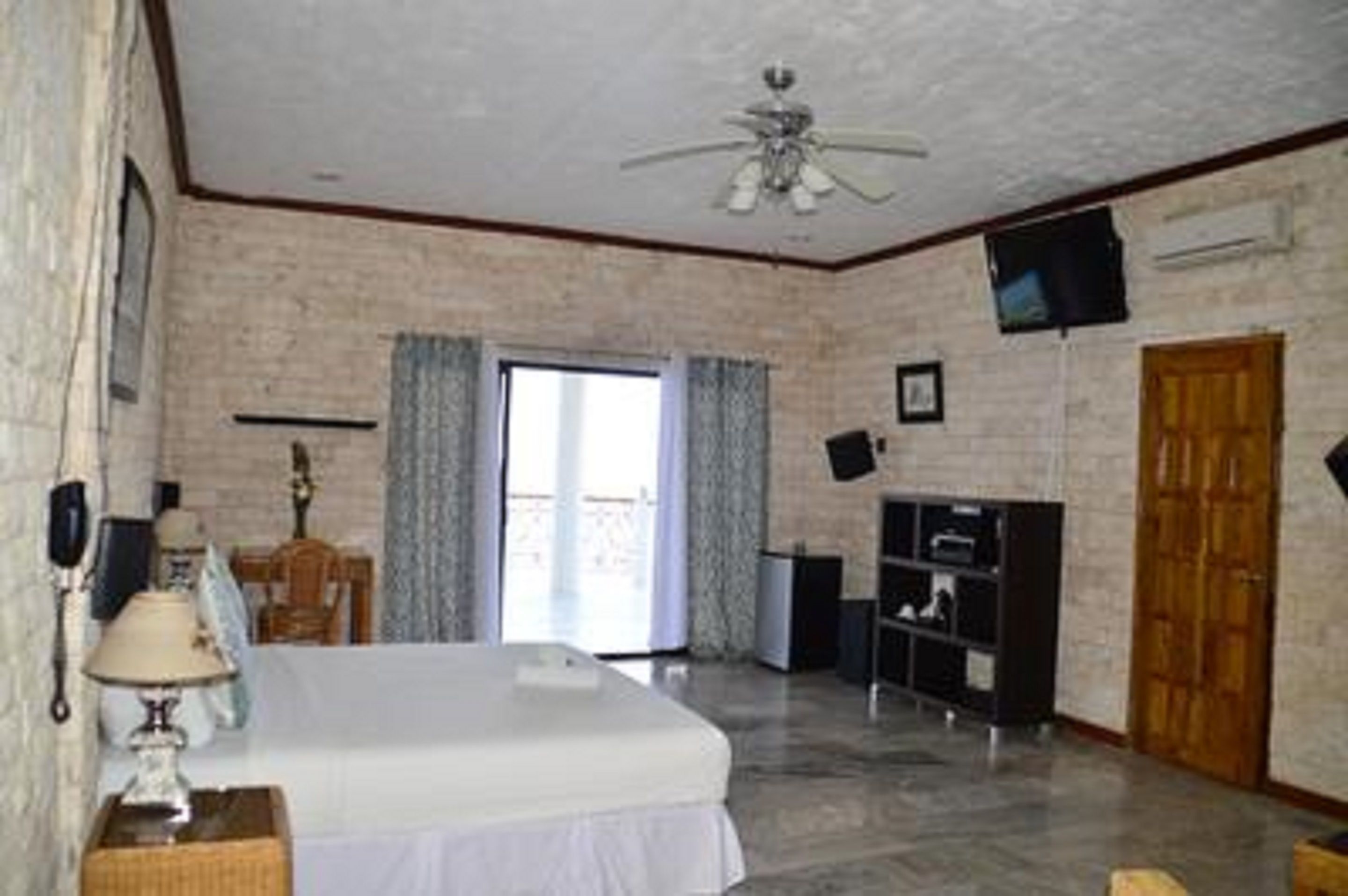A hotel room with a bed, a ceiling fan, a television, a wooden door, and a window with curtains.