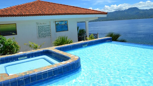 A swimming pool with a tiled edge, a building with a red-tiled roof, and a view of the sea and mountains in the background. - Eden Resort (Santander)