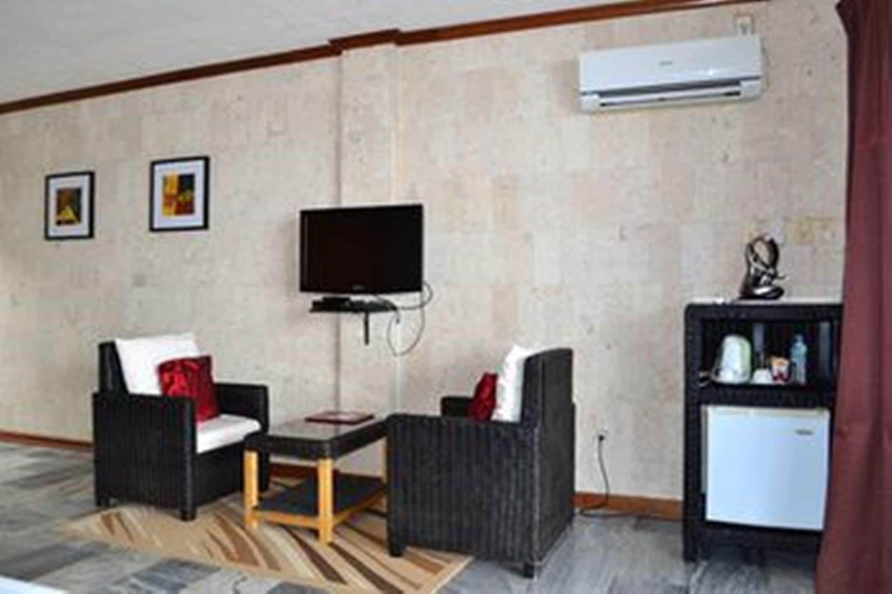 Una habitación con dos sillones negros, una mesita, un televisor de pantalla plana montado en la pared y un refrigerador pequeño a la derecha.