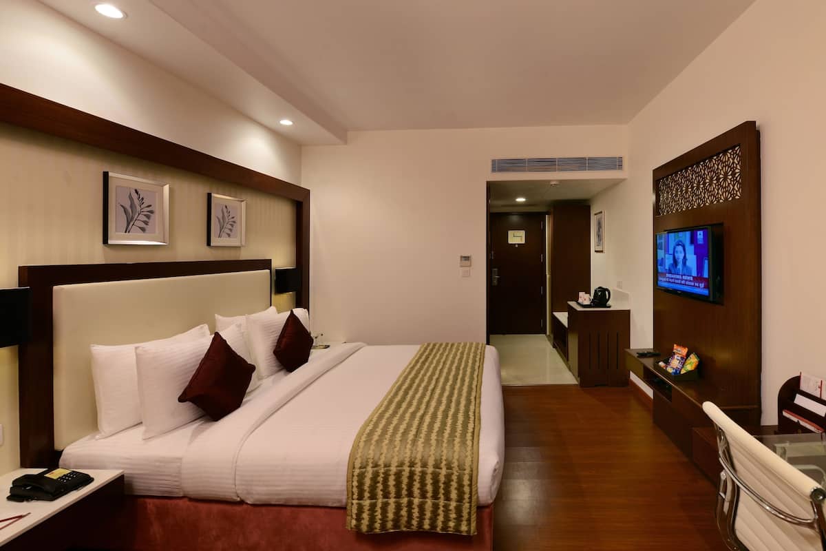 A hotel room with a bed, bedside table, lamp, TV, and a desk with a chair.