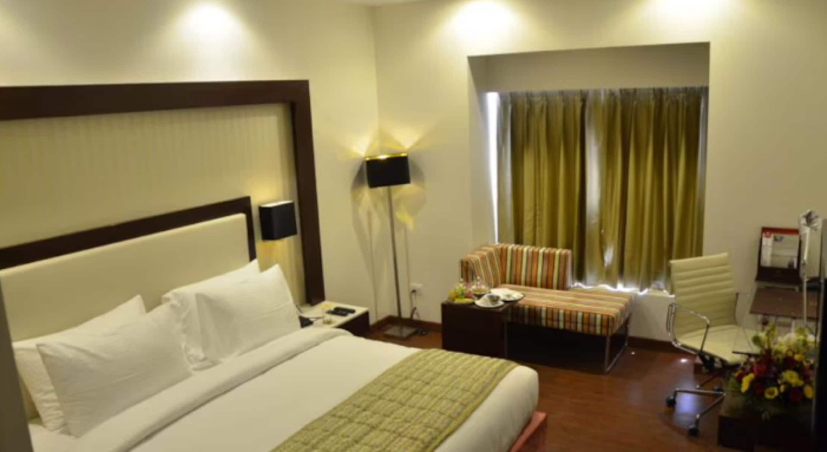 A hotel room with a large bed, a bedside table, a lamp, a chair, a desk, and a window with curtains.