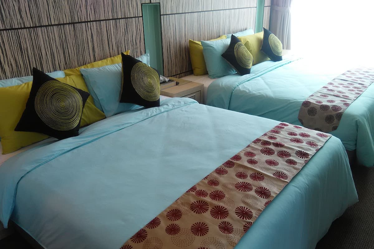 A hotel room with two beds, each with a blue bedspread and a patterned runner. There are multiple pillows in yellow, black, and blue. The room has a striped wallpaper headboard and a green nightstand.