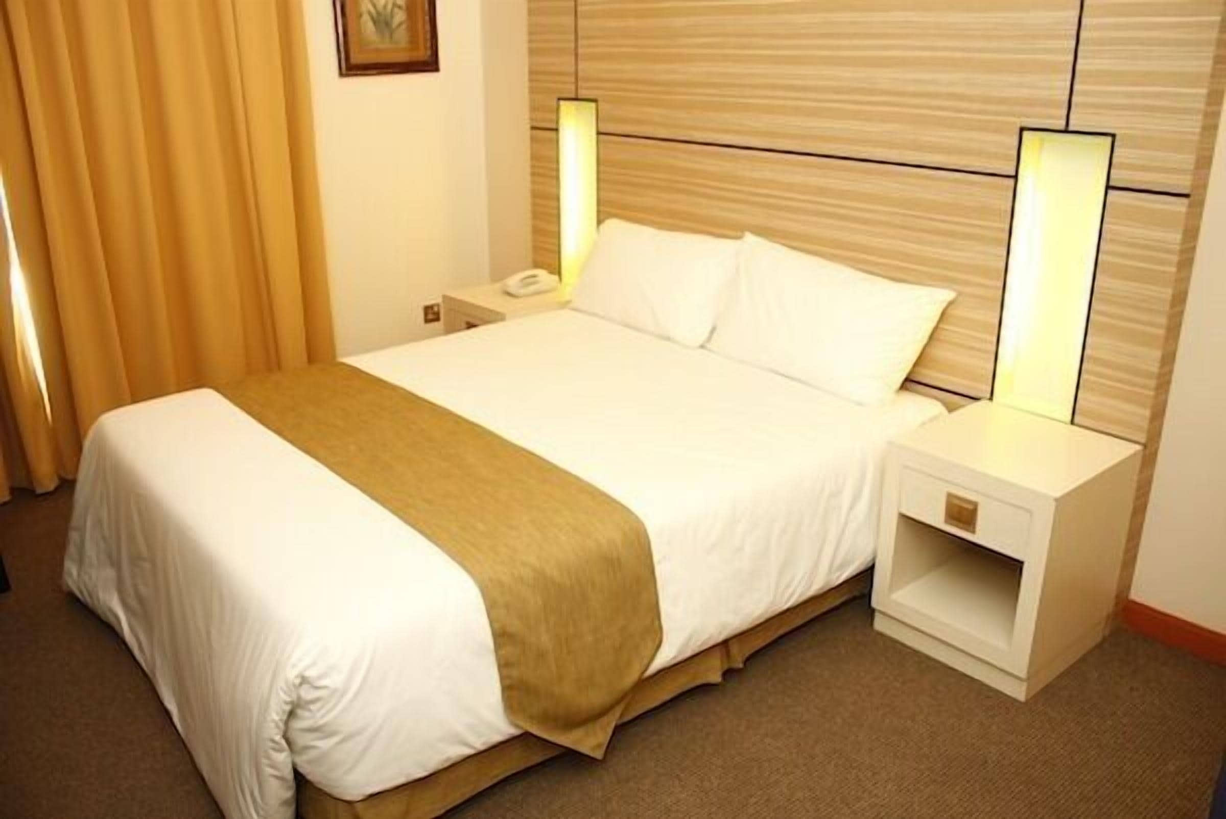 A hotel room with a large bed, white bedding, a wooden headboard, bedside lamps, and a nightstand.