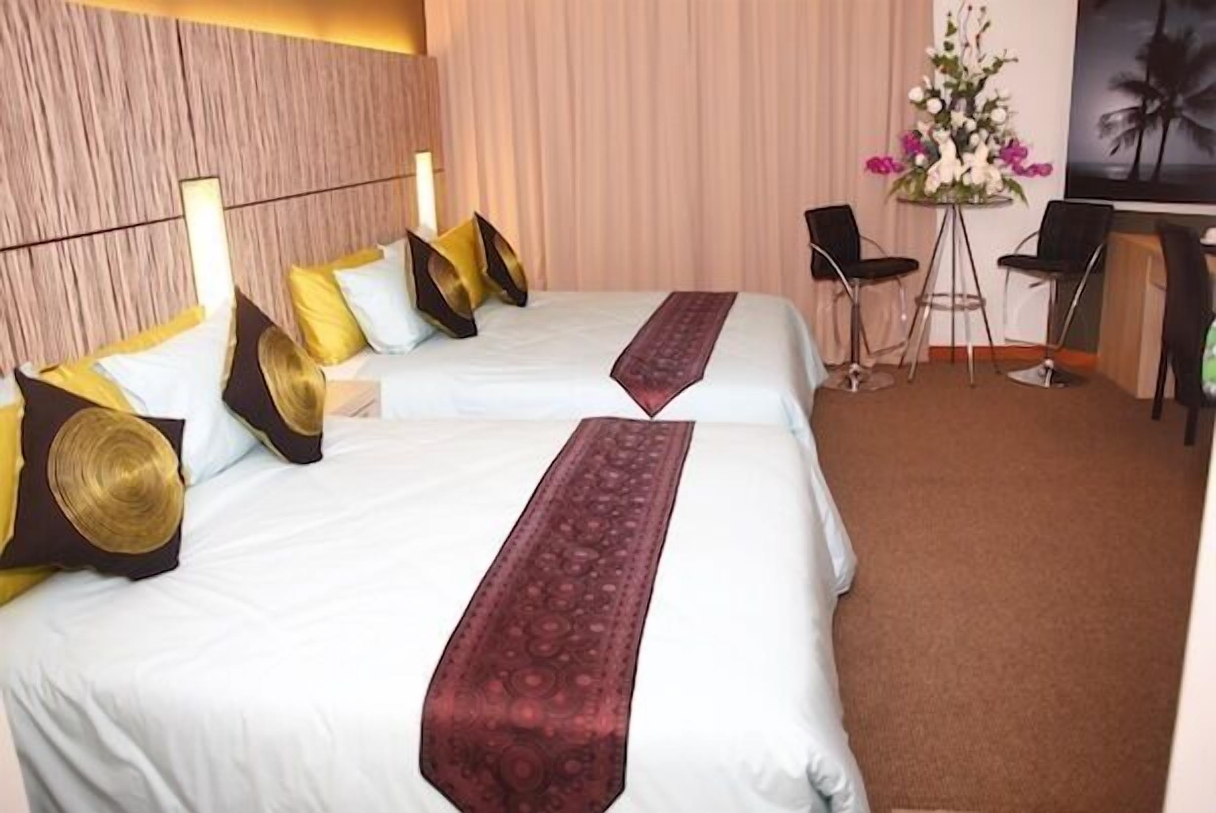 A hotel room with two beds, each with white linens and decorative pillows. There is a desk with two chairs and a vase of flowers.