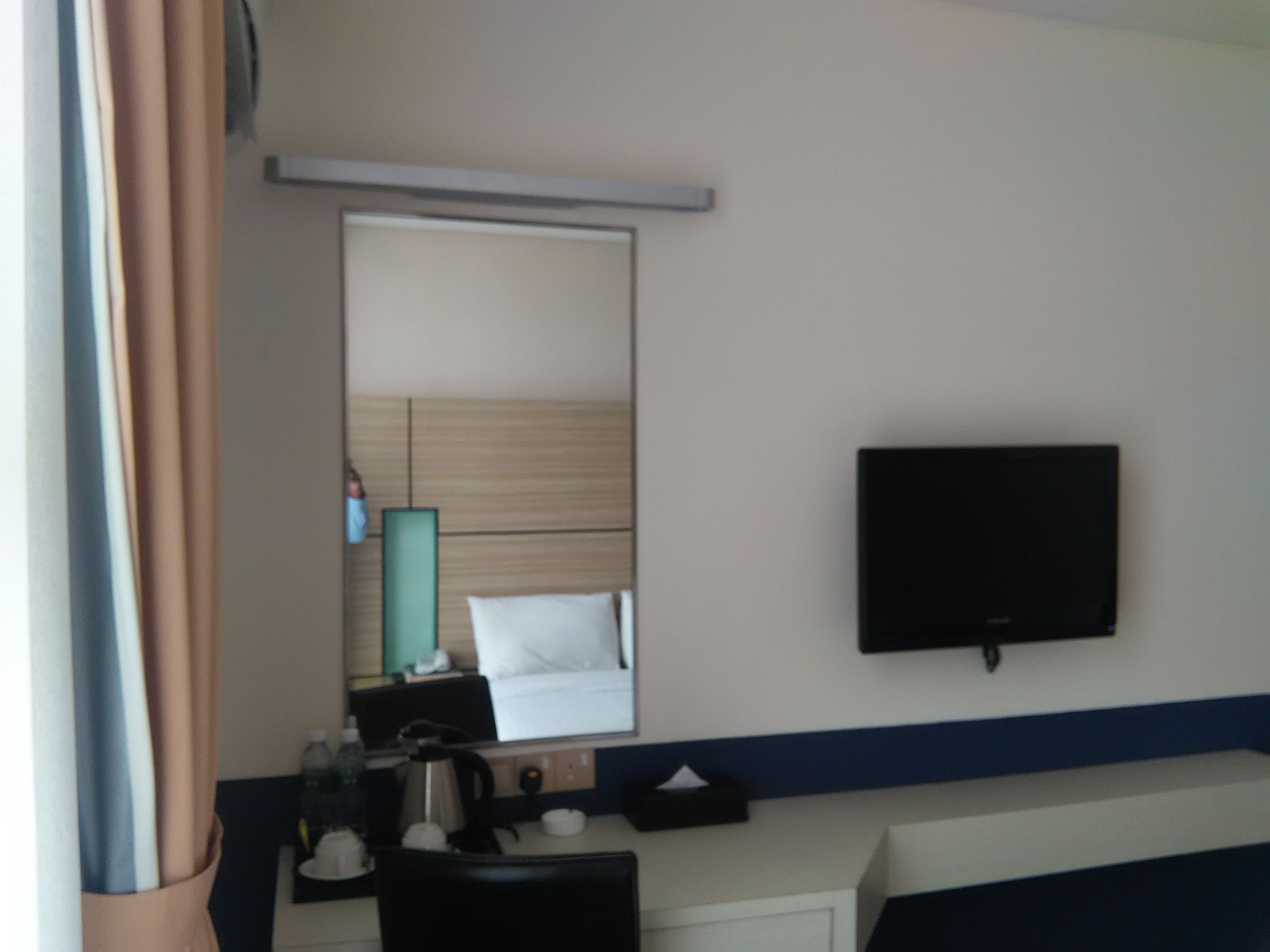 Una habitación de hotel con cama, mesita de noche, espejo, televisor y ventana con cortinas.