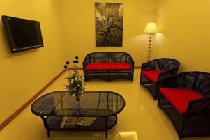 A room with a TV mounted on the wall, a painting above the sofa, a table with a vase of flowers, and a lamp.