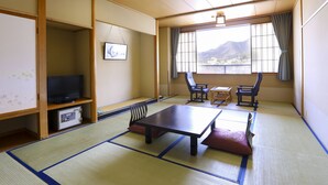 A traditional Japanese room with tatami flooring, a low table, two chairs, a television, and a window with a view of mountains.