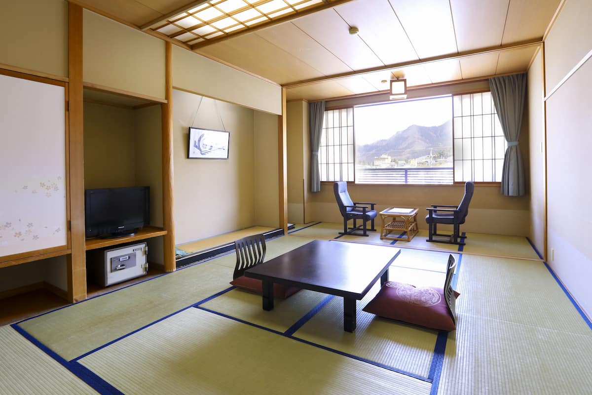 A traditional Japanese room with tatami flooring, a low table, two chairs, a television, and a window with a view of mountains.