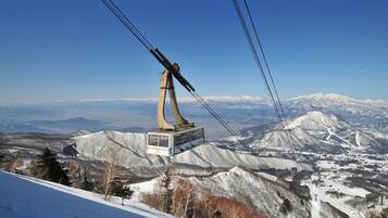 A cable car system with a cabin suspended by cables, set against a backdrop of snow-covered mountains and clear blue sky.