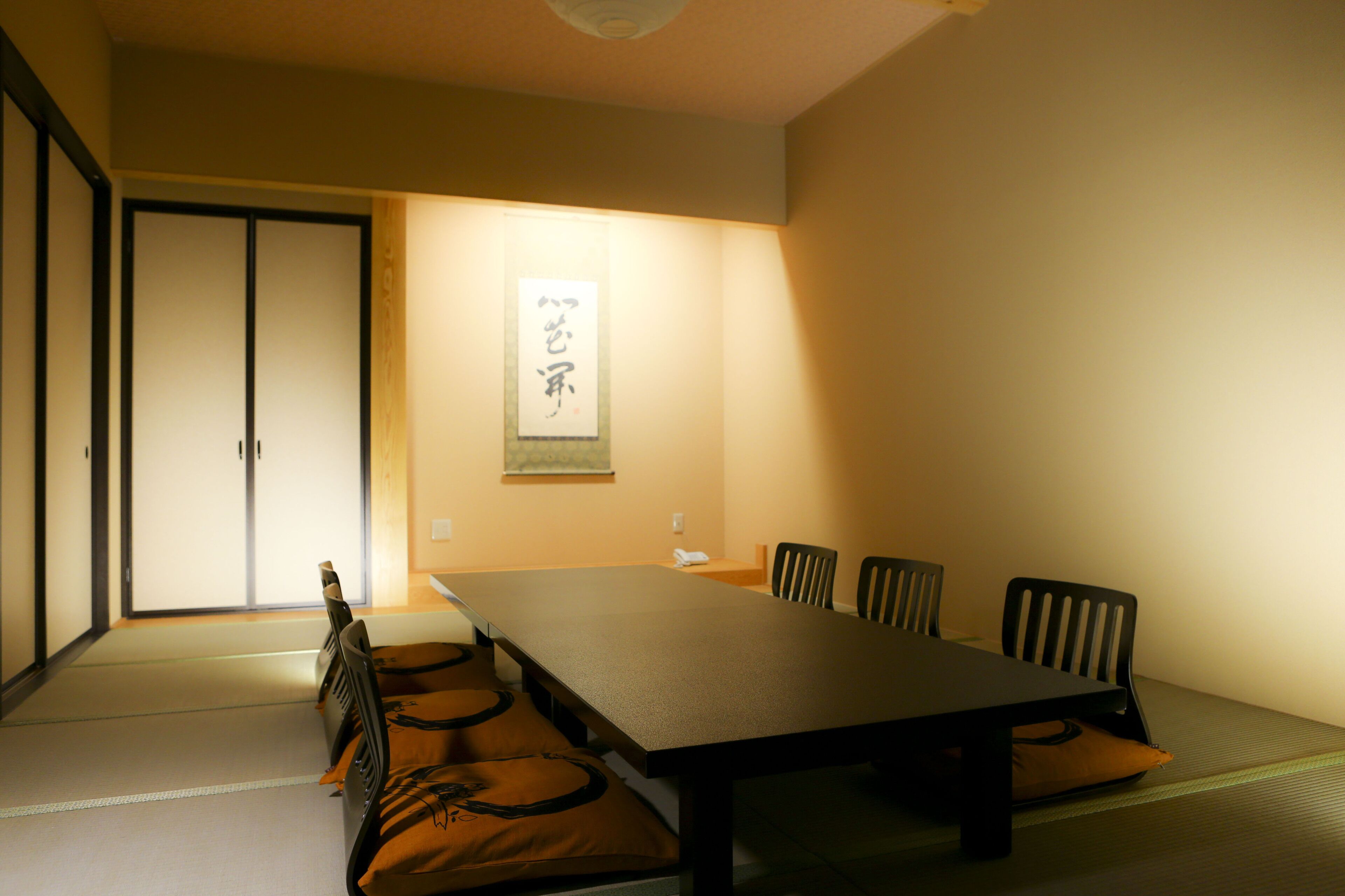 A traditional Japanese room with tatami flooring, a low table, and black chairs.